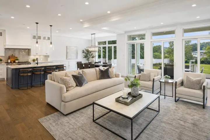Spacious modern living room with beige sofa, large windows opening to a backyard, and a sleek kitchen and dining area.