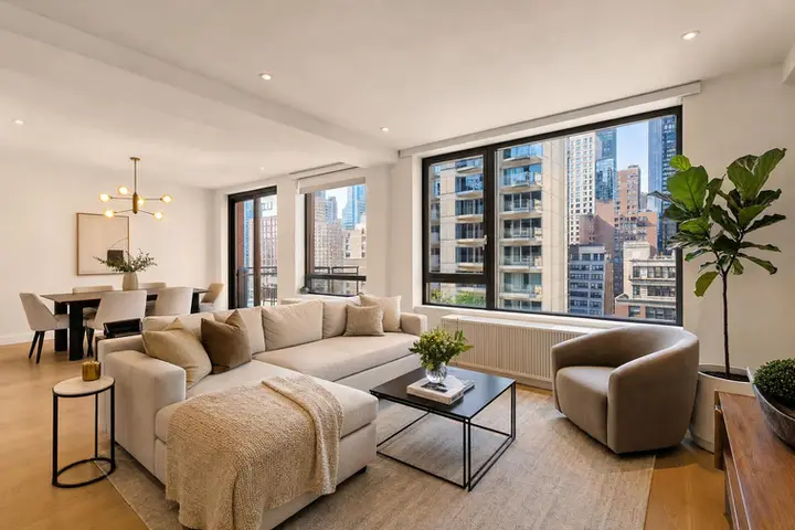Modern living room with large windows showcasing an NYC skyline, beige sectional, and dining area with pendant lighting.