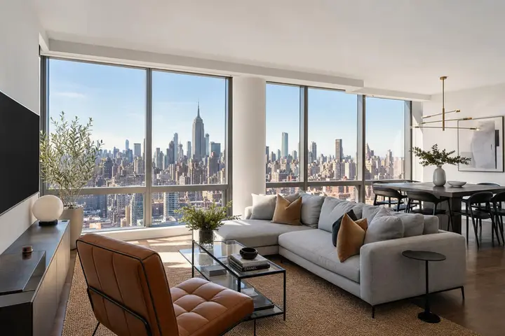 Modern living room with large windows displaying NYC skyline, featuring a cozy sofa and dining area with stylish decor.