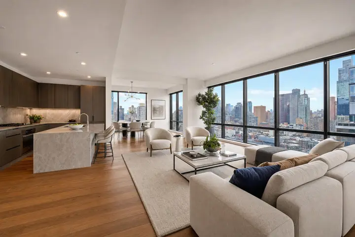 Modern open-plan living room with beige sofas, large windows offering NYC skyline views, adjacent to a sleek kitchen.