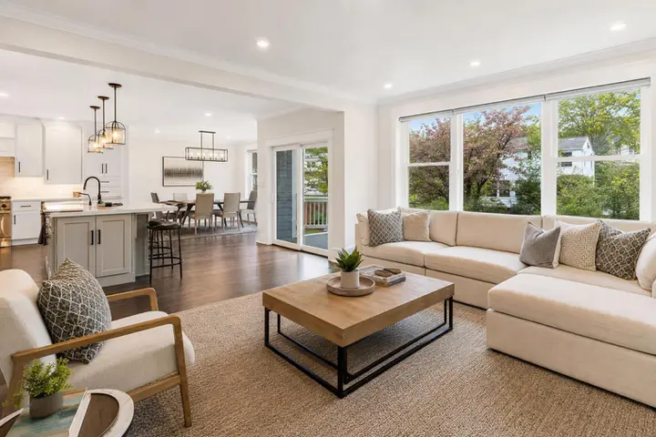 Open-concept living room with beige sectional, wooden coffee table, and view of dining area and kitchen in bright space.